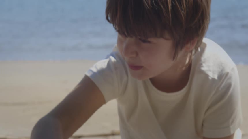 Close-up of a contemplative young boy playing on a sandy beach with the ocean in the background