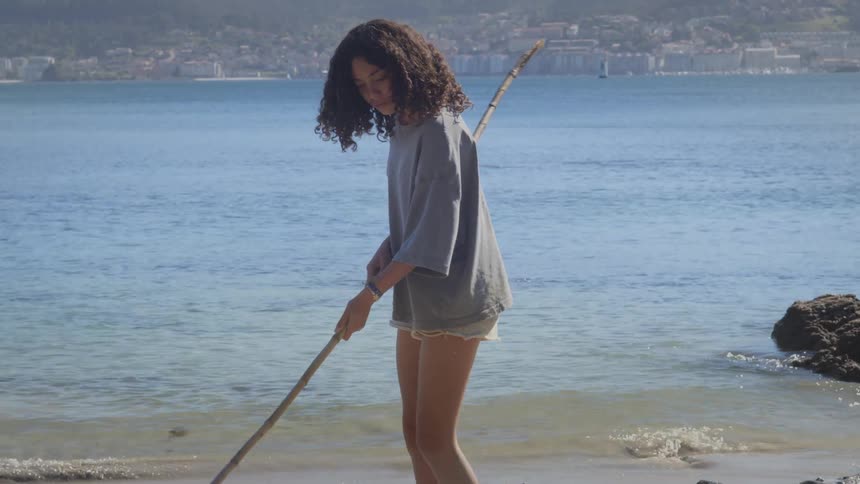 A young woman with curly hair draws in the sand with a long stick as gentle waves lap at her feet on a sunny beach