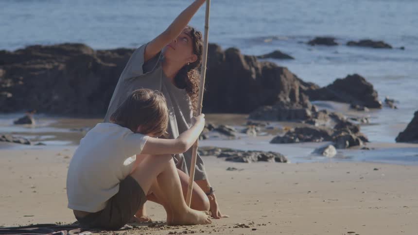 A young boy and his older sister spend a sunny day at the beach, playing together with a bamboo stick in the sand