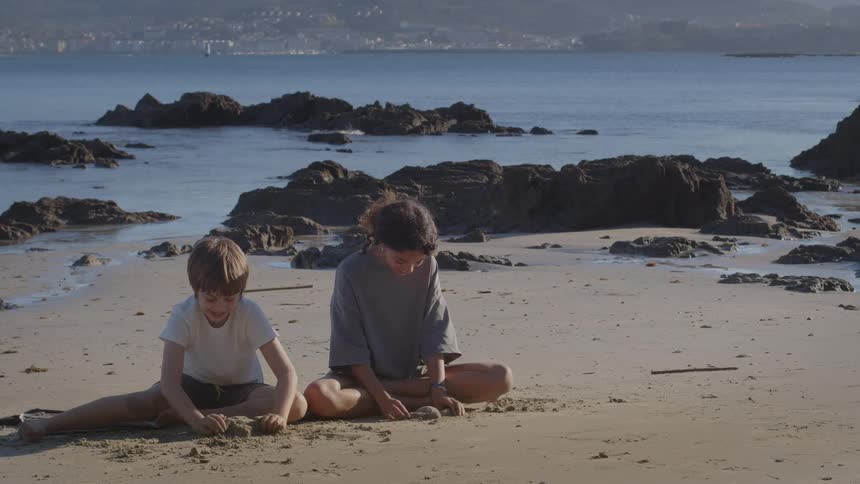 A young boy and girl sit together on a quiet, sandy beach, happily playing in the sand