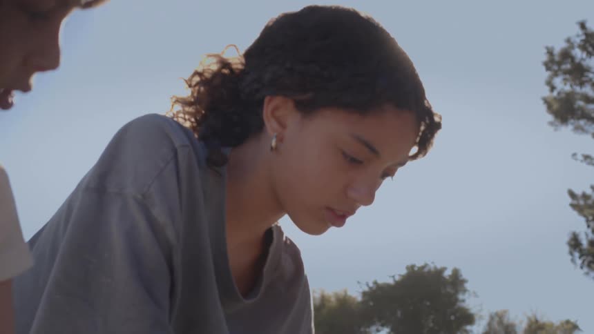 A low-angle, slow-motion shot of two young people focusing on a task outdoors with the sun creating a lens flare