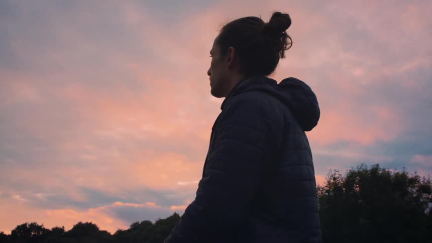 A man with his hair in a bun peacefully observes the stunning pink and orange colors of the sunset sky