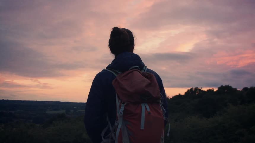 A lone hiker stands on a hilltop, taking in the breathtaking view of a pink and purple sunset over the countryside