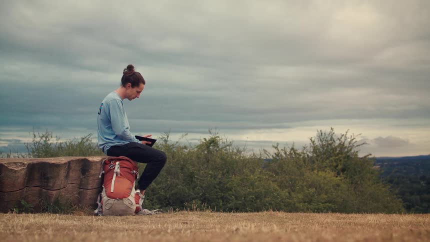 A young hiker with a backpack sits on a tree stump on a hilltop, sketching on a tablet under a vast, cloudy sky