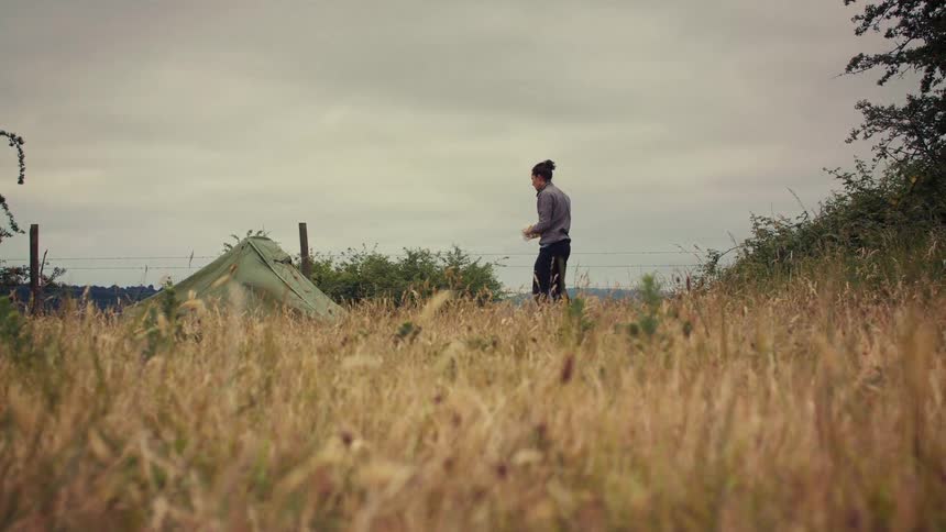 A person walks through a grassy field next to their green tent on a cloudy, overcast day