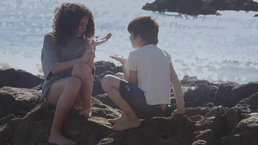 A young boy and girl sit together on coastal rocks, curiously examining their beach discoveries in the bright sunlight