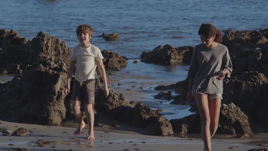 A young boy and a girl in slow motion jump from the coastal rocks onto the wet sand to explore the shoreline