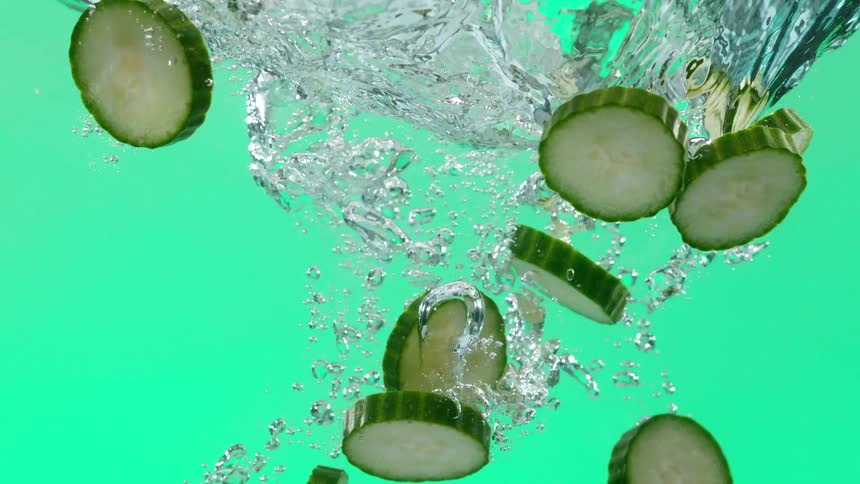 Slices of fresh cucumber splash into water in slow motion against a green background, creating bubbles