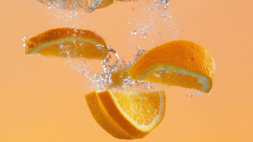 Fresh orange slices gracefully fall into water, creating bubbles in slow motion against a matching orange background