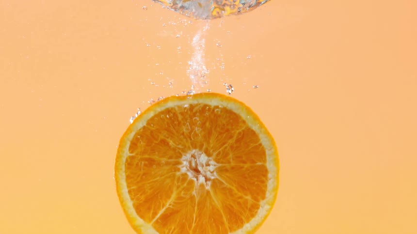 A vibrant slice of orange slowly sinks into water, leaving a trail of sparkling bubbles against an orange background
