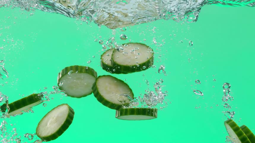 Super slow motion of fresh cucumber slices falling into water against a vibrant green background