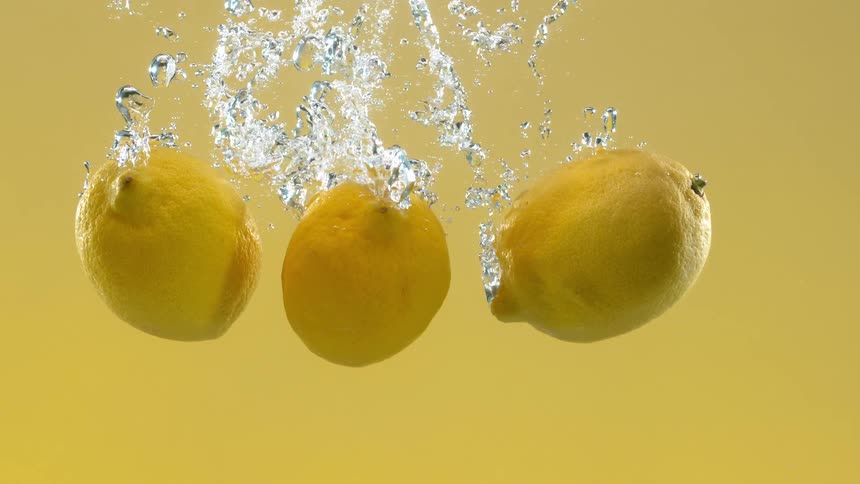 Three whole lemons splash into water against a yellow background in slow motion
