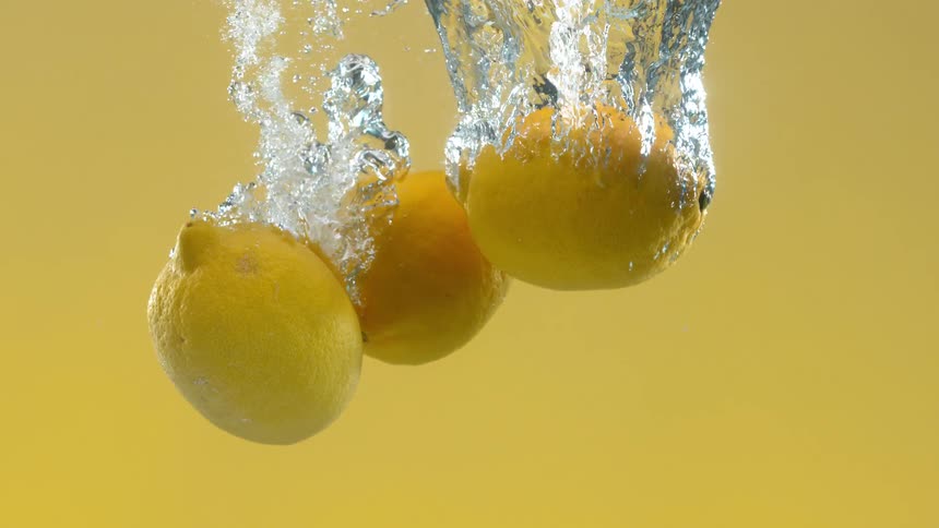 Three fresh yellow lemons fall into clear water in slow motion, creating a splash and bubbles against a yellow background