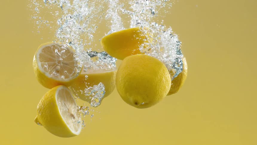 Cut and whole lemons drop into clear water against a yellow background in super slow motion