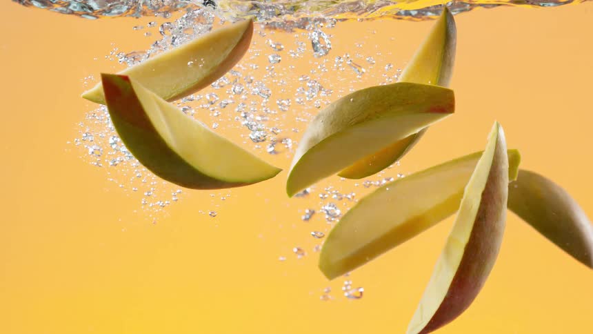 Fresh mango slices make a refreshing splash as they fall into water in super slow motion against a yellow background