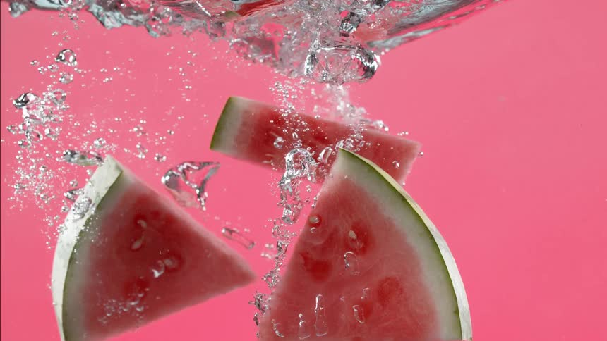 Fresh watermelon slices fall into water with a splash against a vibrant pink background in slow motion