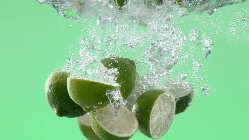 Fresh halved limes splash into clear water in slow motion, creating a burst of bubbles against a vibrant green background