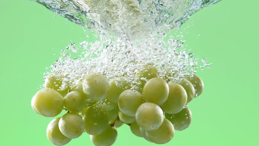 A lush bunch of green grapes plunges into clear water, creating a cascade of bubbles in super slow motion against a vibrant green screen
