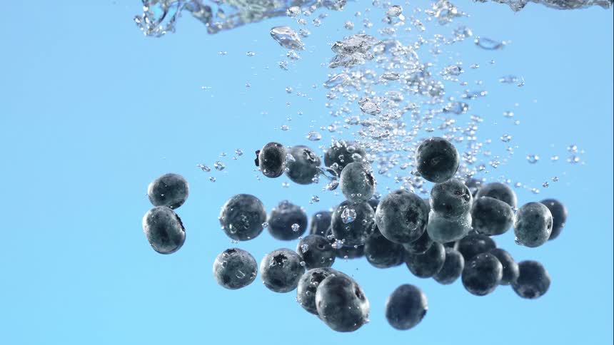 A handful of fresh blueberries plunges into clear water, creating a cascade of bubbles in a super slow-motion shot against a vibrant blue backdrop