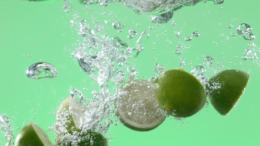 Fresh lime halves splash into bubbly water in super slow motion against a vibrant green background