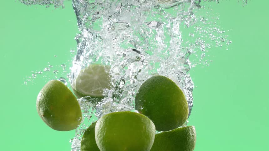 Fresh sliced limes fall into sparkling water, creating beautiful bubbles in super slow motion on a green screen background