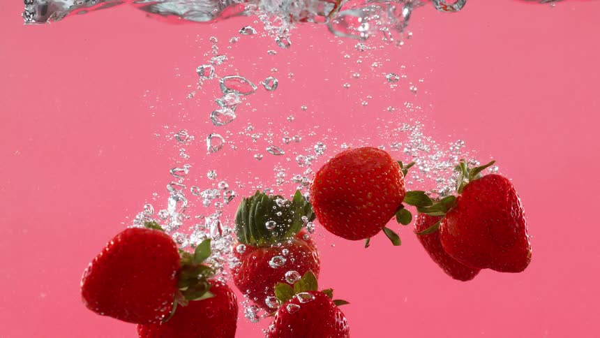 A handful of bright red strawberries splash into clear water against a pink background in super slow motion