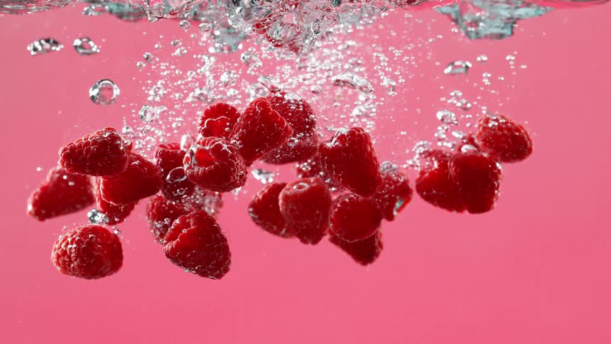 A handful of fresh red raspberries fall into clear water, creating a cascade of bubbles and splashes in super slow motion against a vibrant pink background
