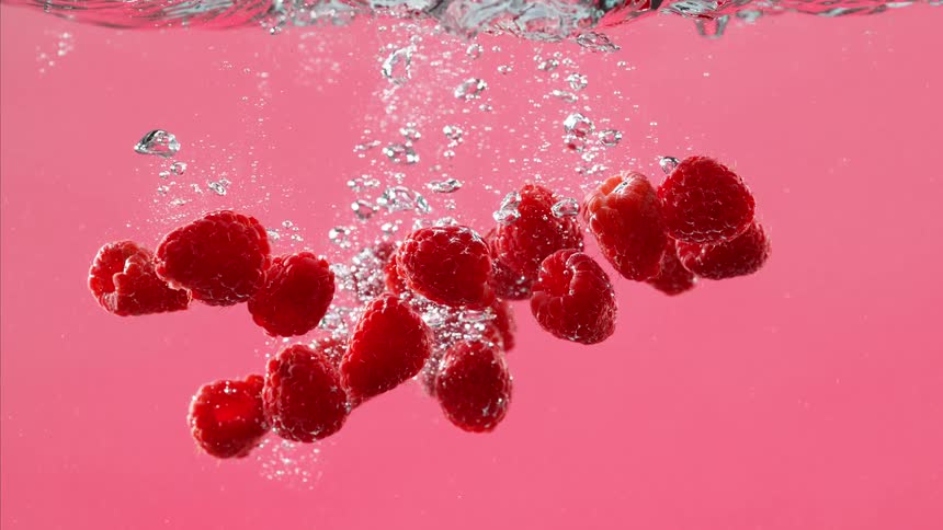 Fresh raspberries plunge into sparkling water against a vibrant pink background in an ultra slow motion shot