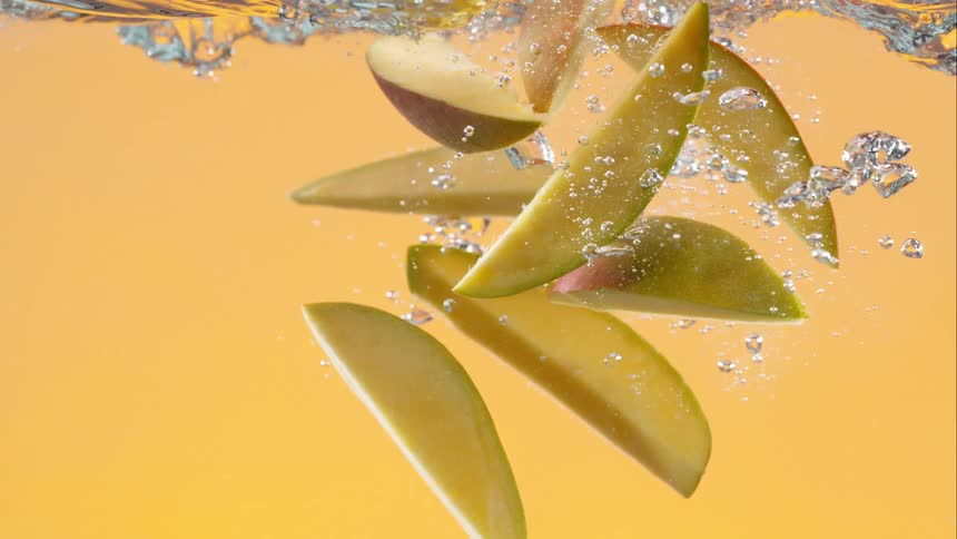 Fresh mango slices fall into water with a splash in super slow motion against a vibrant yellow background