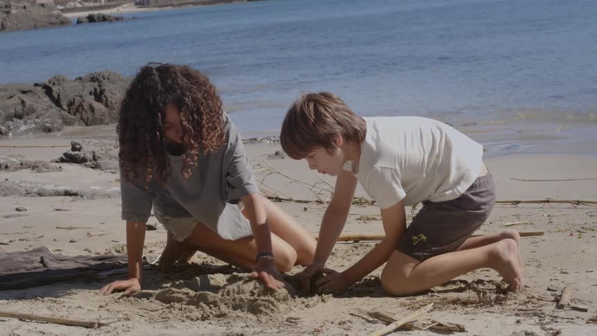 A young boy and girl kneel on the sand as they play together at the beach on a sunny day