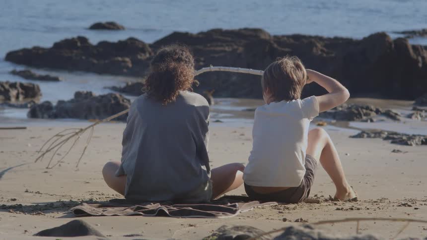 Two children sit together on a towel at the beach, peacefully gazing at the ocean