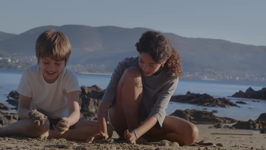 Kids play with sand together on a rocky beach with calm ocean and mountains in the background