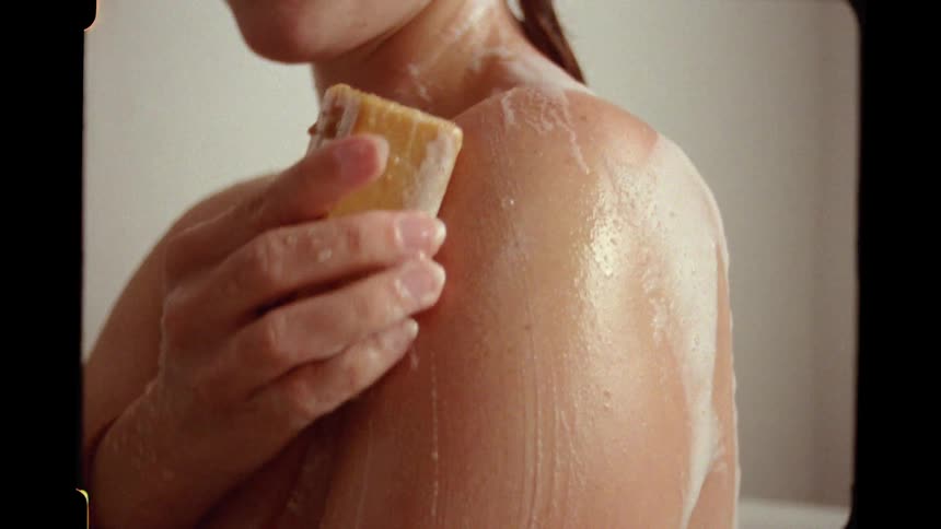 A close-up shot of a woman's shoulder as she lathers her skin with a bar of soap in the shower, with a vintage Super 8 film aesthetic