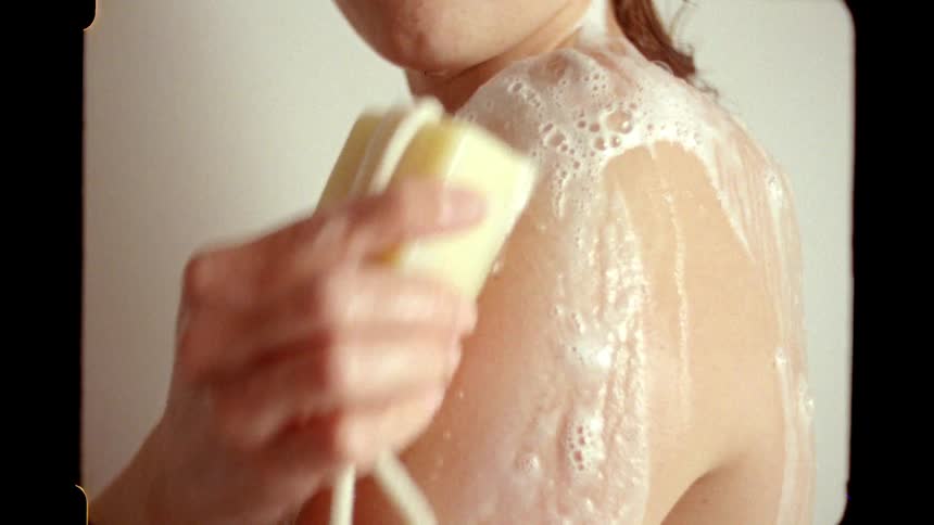 A close-up shot of a woman lathering her sudsy shoulder with a bar of soap in the shower, captured with a vintage film aesthetic