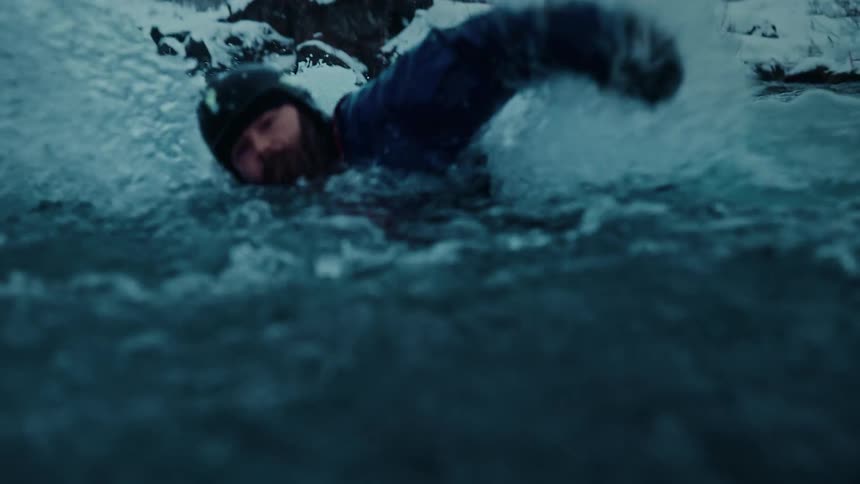 A determined swimmer in a wetsuit battles the powerful current of an icy river in a dramatic slow-motion shot through a snowy landscape