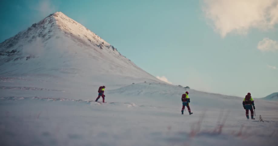 A team of three hikers with backpacks and poles brave the wind as they traverse a vast, snowy mountain landscape