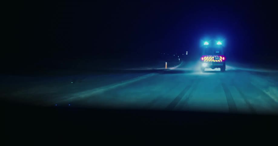 POV shot following an emergency rescue vehicle with blue lights flashing down a dark, snow-covered road at night