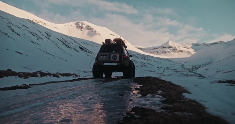 A rugged off-road vehicle with large tires drives down an icy road into a vast, snow-covered mountain valley during sunset