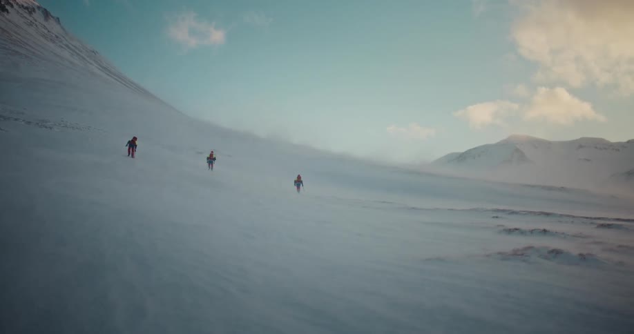 A group of hikers with backpacks ascend a vast, snow-covered mountain during a windy blizzard