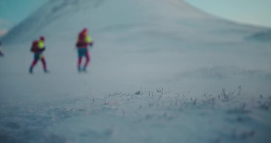A low-angle, out-of-focus shot shows three hikers with backpacks trekking across a vast, snowy mountain landscape during a windy winter day