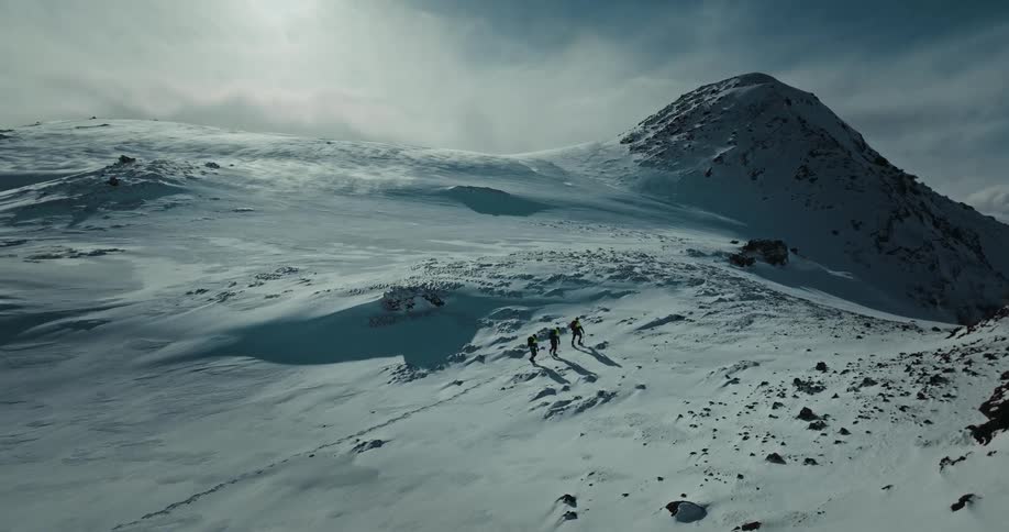An epic aerial view follows a team of mountaineers as they trek up a vast, sunlit, snow-covered mountain