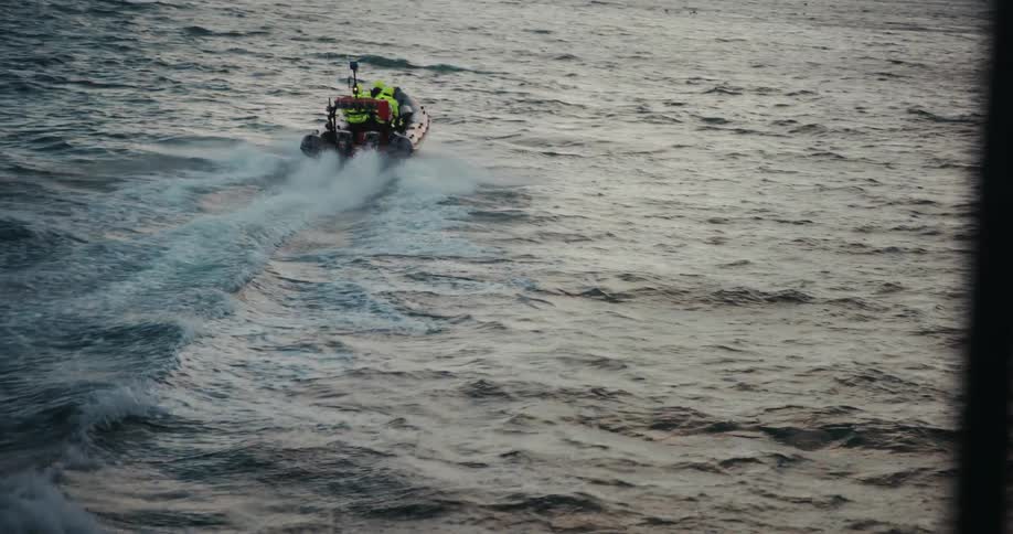 A rigid inflatable boat with a crew in bright safety gear speeds across the cold, choppy sea towards a distant snowy coastline