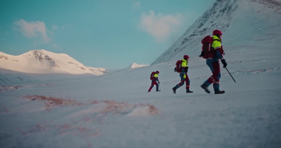A team of three mountain rescuers equipped with backpacks and helmets hikes through a vast, snow-covered valley in slow motion