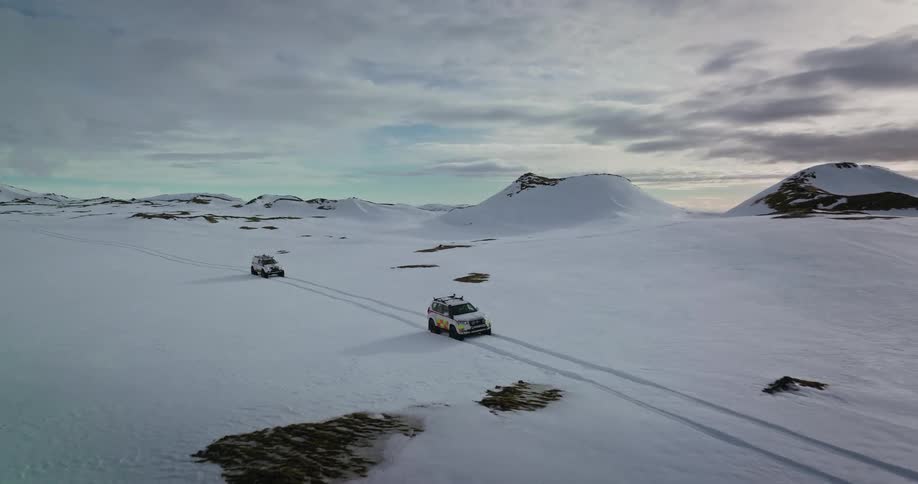 Aerial view of two expedition SUVs driving in tandem across a vast, snow-covered landscape in the mountains