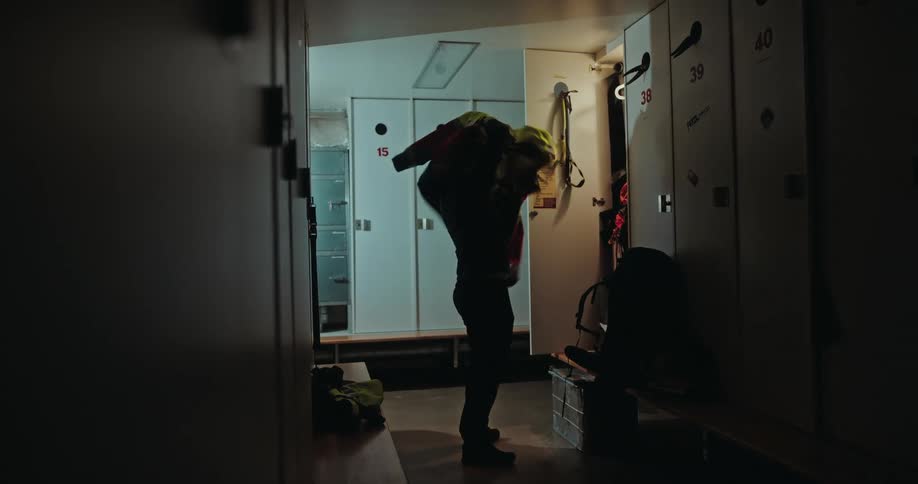 A man gets ready in a dark locker room, putting on his high-visibility jacket and large backpack before his expedition