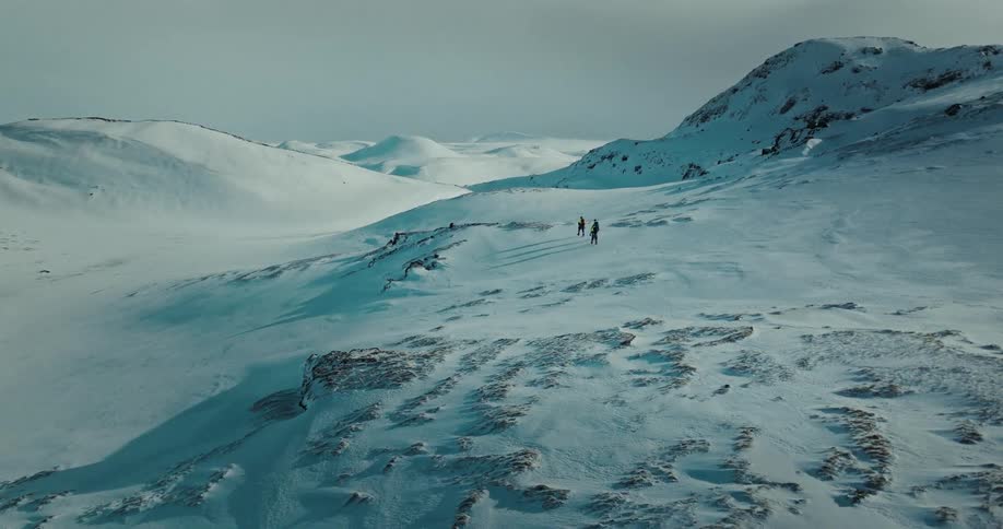 An expansive aerial view shows two hikers trekking across a desolate and seemingly endless snow-covered mountain range