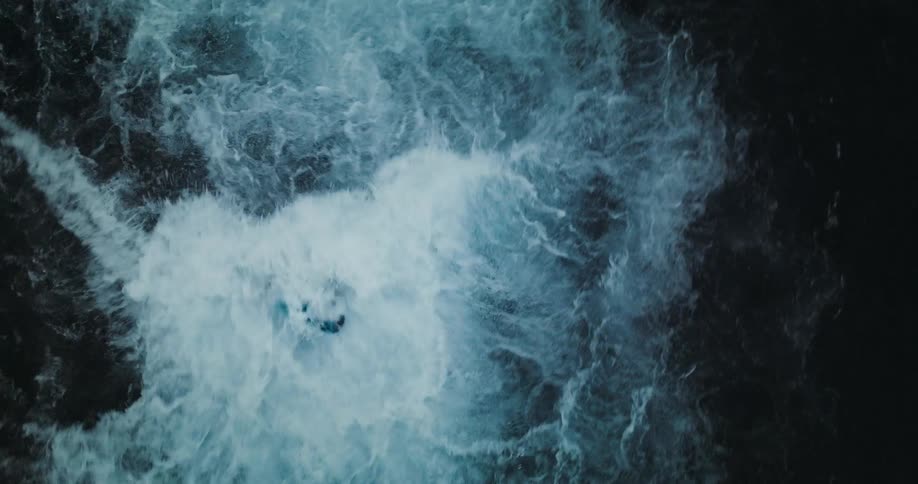 Top-down aerial slow-motion shot of an extreme athlete jumping into a powerful, icy river and navigating the turbulent rapids