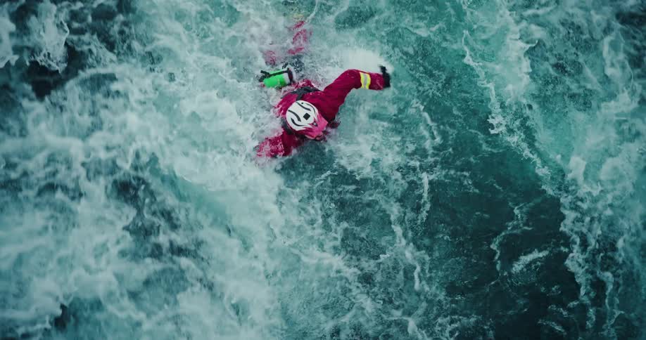 A high-angle, slow-motion shot shows a rescue swimmer diving headfirst from a rock into a powerful, turbulent river during a training exercise