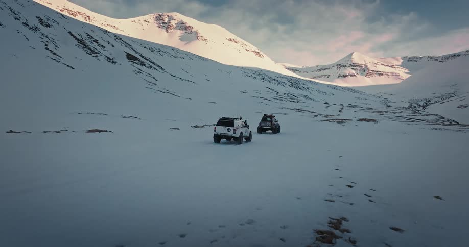 An epic aerial shot follows two rugged expedition vehicles as they drive away through a vast, snow-covered mountain pass at sunset