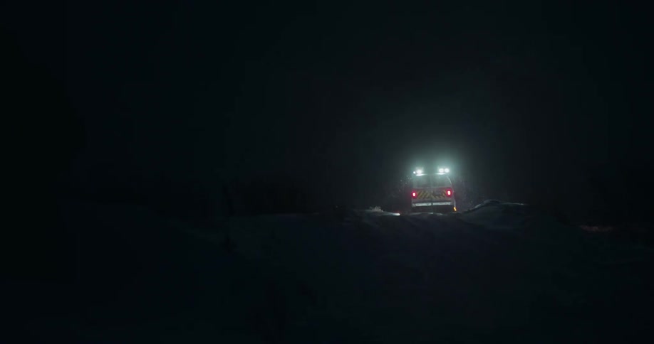 A search and rescue van drives away from the camera into a dark and snowy landscape during a heavy snowstorm at night